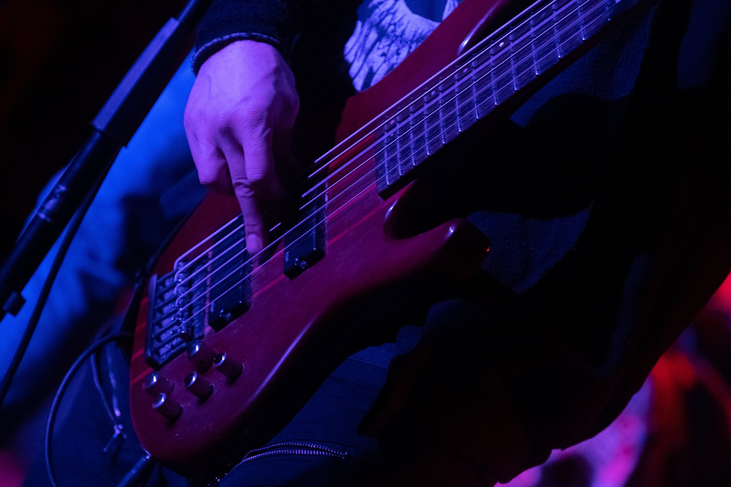 Close-up of a person's hand playing a red electric bass guitar on stage with stage lighting in the background.